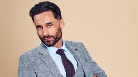 Luke Harden, dressed in a grey suit and purple tie in a modelling shot