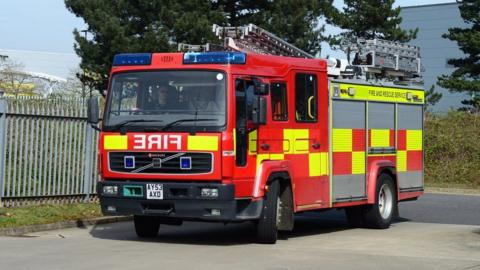 A fire engine is pictured driving. The red and yellow vehicle has blue lights on it as well as firefighting equipment including ladders resting on its roof.