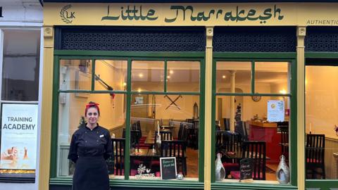 A woman in a black uniform stands smiling outside a restaurant which has a sign saying 'Little Marrakesh'.