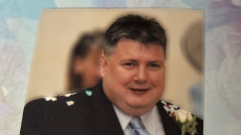 A wedding photograph of John Smyth wearing a black suit and blue-grey tie. He has closely cropped, dark hair. Confetti can be seen on his shoulders and he wears a yellow flower in his lapel.