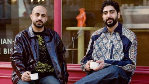 The two owners of the cafe holding coffee cups outside a red-painted cafe
