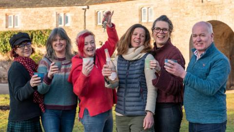 Five women and a man smiling into the camera holding mugs. The woman on the centre-right is holding a bottle of fizzy wine and the woman on the centre-left is holding some keys above her head.