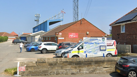 A carpark in front of a small football stadium with a blue covered stand. A sign in the car park says: Welcome to Whitby Town F.C.