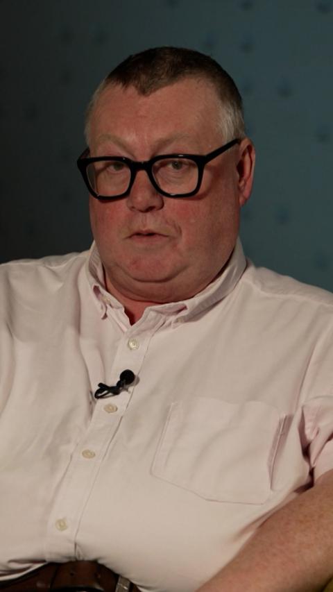 A man with short grey hair and black glasses sits in front of a greyish blue wall. He is wearing a light pink shirt and has a black lapel mic attached.