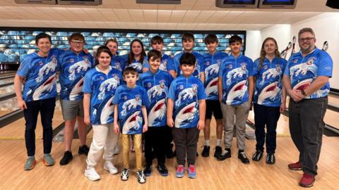 A group of 13 young bowlers alongside their coach. They are posing for a group photo in a bowling alley and smiling at the camera. They are all wearing matching blue shirts which depict a shark jumping out of waves. 