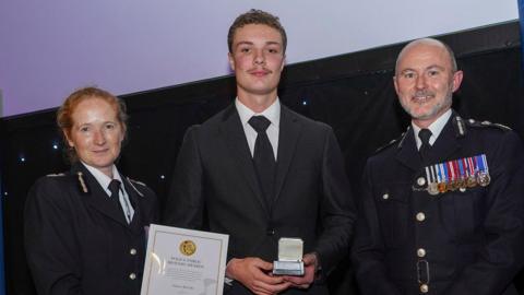 Ashton McCabe standing in the middle holding a small silver plaque. He is wearing a black suit and tie. A woman on his right is handing him a certificate. There is a man on his left who is wearing a row of medals on his chest.