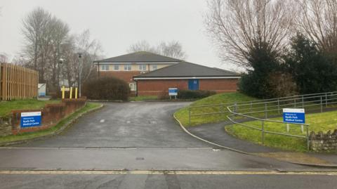 The driveway leading up to Ryalls Park Medical Centre in Yeovil. There are blue signs either side of the driveway, with a brick wall and grass verge. The red brick GP surgery building is in the background, surrounded by trees. The sky is dull and grey.