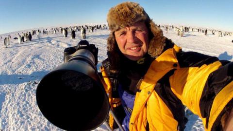 a man in a yellow jacket and furry hat with a large camera 
