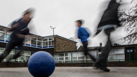Three children in trousers are playing with a blue football. They are blurred and a school building is in the background.