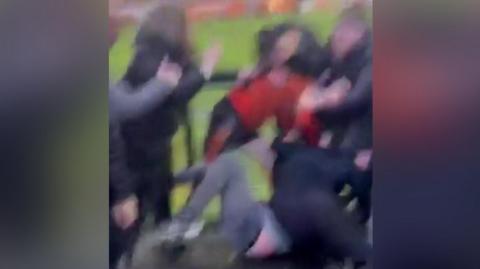 Several people including what looks like a player are brawling and fighting by the side of the pitch. Four or five people are grappling one another with two people stood on the left remonstrating with those involved.