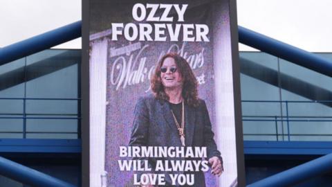 A board that has a picture of Ozzy Osbourne on it. It reads "Ozzy forever, Birmingham will always love you"