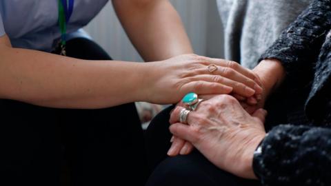 Two people holding hands. Only their forearms and hands are visible. The arm on the left belongs to a younger woman who has a diamond ring on her finger. The arm on the right of the image is covered by a dark knitted top and the person wearing it also has two rings on their fingers.