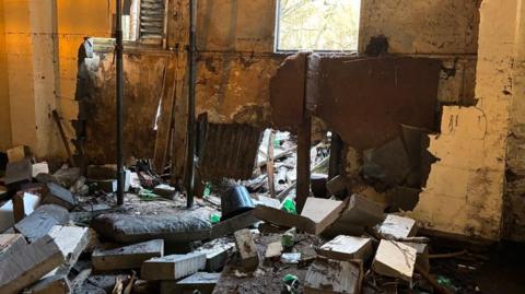 A pile of rubble including bricks, a plastic bucket and chunks of wood lies strewn across the floor of an industrial unit. A wall has been knocked through underneath a window, with daylight coming through. 