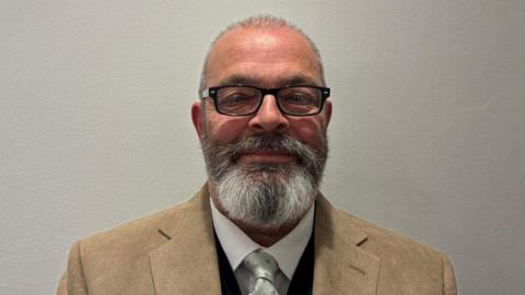 A man with glasses and shaved head and beard wearing a light brown jacket and shirt and tie