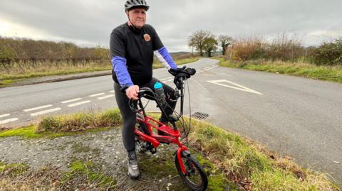 Tim Bright is dressed in black Lycra, with purple sleeves showing. He is stationary, sitting on a red Raleigh Chopper, on the verge, at the junction of two roads. He is wearing a cycle helmet, and a bottle of water is in the handlebars. Trees and hedgerows line the roads.