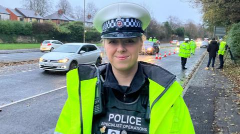 PC Ella Thornton is pictured standing in front of a road. The first lane is coned off with police officers directing traffic. PC Thornton has blonde hair and she is smiling. She is wearing a police hat and a fluorescent jacket with police uniform and a radio underneath.