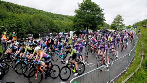 Professional cyclists begin a road race in a rural location. The road s flanked by silver fencing, trees and greenery.