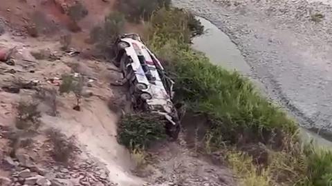 A frame from a video showing a wrecked passenger bus at the bottom of a deep ravine. The terrain is rocky and sandy with a shallow river snaking across the bottom of the ravine. The bus looks utterly destroyed.
