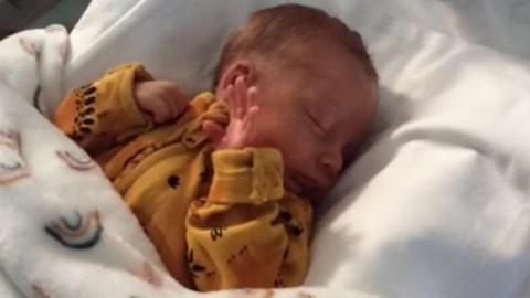 A newborn baby wearing a yellow sleep suit, laying in a bed covered with a blanket.