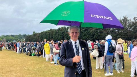 A man with grey hair wearing a navy blue suit and a striped white and navy blue tie is holding an umbrella. He is looking at the camera. Behind him, in a field, are queues of people, waiting to go into Wimbledon.