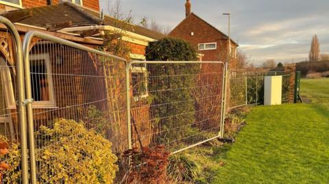 Heras fencing installed on the very edge of residents' homes. Their back gardens are on the left of the image, inches away from the fencing which blocks the view from their windows. On the right side of the fencing is a lush green field. 