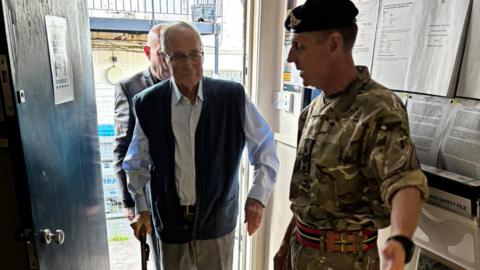 An elderly man, wearing a pale blue shirt and a dark-coloured gilet, is seen entering a building. A man in military uniform is seen offering him directions.