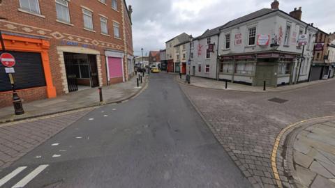 West Row is a street lined with shopfronts. A pub can be seen on the left. Side roads coming off West Row are cobbled.