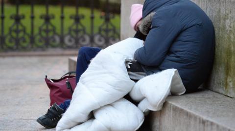 A person wrapped in a duvet is falling asleep on some stone steps in a park. They are wearing a padded dark blue jacket and a pink woolly hat.