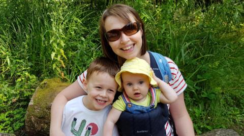 Nicola Wheatley is sitting on a  rock in a green space with her children on her lap. She and Oscar are smiling at the camera and baby Ffion is wearing a yellow summer hat and dungarees.