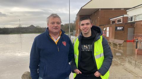 Two men in warm clothes stand outside of a brick building. There is a flooded Rugby pitch to their left.