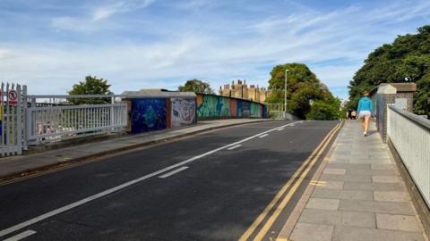 Mill Road bridge in Cambridge with a woman walking on the pavement. She is wearing shorts and a blue top