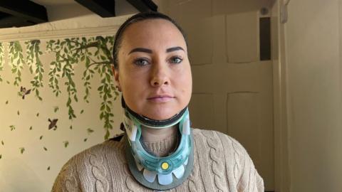 Ball is photographed looking into the camera with a neutral expression. She is wearing a beige cable knit jumper, and has her dark hair tied back in a ponytail. She is also wearing a translucent, light blue neck brace. Ball is stood by a window, with natural light hitting her face. The room she is stood in has white walls, with dark beams on the ceiling. A mural on the wall behind her has a tree with trailing leaves and two small images of birds.