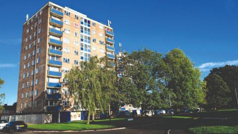 Seagry Court, a high rise block with green trees surrounding it. The sky behind it is blue.