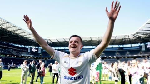 Joe Tomlinson standing looking at a home crowd with both arms aloft and huge smile, wearing MK Dons home kit