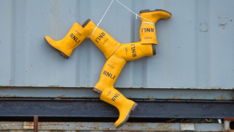 An Isle of Man triskelion made of several pairs of yellow RNLI wellies is hanging from the wall of a shipping container
