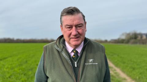 Colin Rayner, a farmer based in Berkshire and Surrey, looks straight at the camera. He has dark greying hair, and is wearing a pink shirt, green and yellow tie, green zip up top and a green gilet. Behind him is a green field and a grey sky