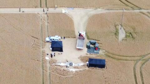 A drone image of investigators at the scene of the crash. A white plane can be seen badly damaged and there are several tents set up around it. It is all based in a large field.