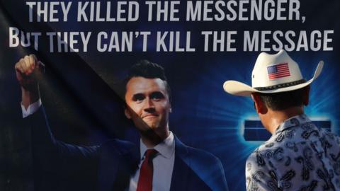 A man wearing a cowboy hat with a US flag on the back looks at a banner with a large picture of Charlie Kirk on it - the message on the banner reads "they killed the messenger, but they can't kill the message"