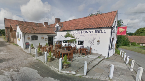 Exterior of the white pub with 'The Hunny Bell' written on the side and patio tables and chairs on a slabs of paving