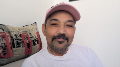 A young man wearing a white t-shirt and pink baseball hat seated indoors next to a decorative cushion featuring iconic red London telephone boxes.
