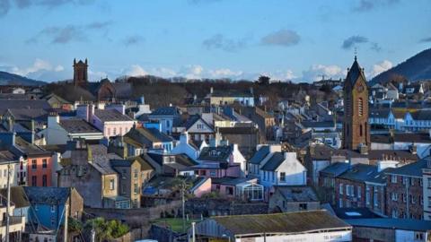 The colourful rooftops of buildings and homes in Peel, a church spire jumps out of the skyline on a bright day with blue skies.