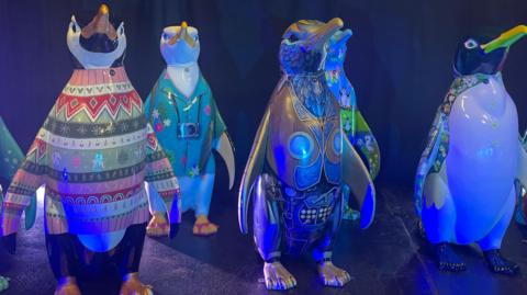 A waddle of penguins stand on a stage with a black floor and curtained black background. The penguins are dressed in multi coloured coats and jumpers and have gold, yellow and green beaks. 