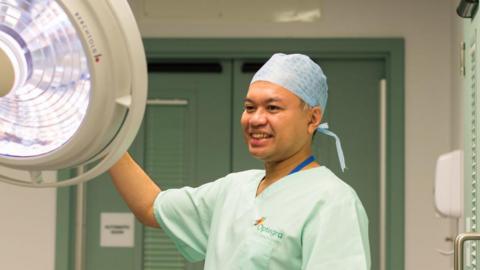 A man wearing medical scrubs in pale green, looking towards a large operating light.