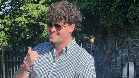 A headshot of a man with sunglasses on and a white, purple and green checked shirt. He smiles and is looking slightly off camera. He has his thumb up.