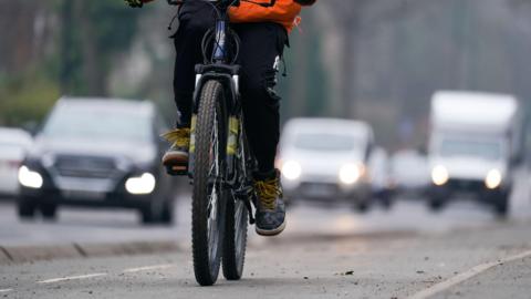A  woman rides a bike along a cycle lane. The cyclist is pedalling. A number of cars are in the background with their lights on. 
