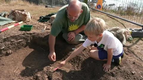 The picture shows an archaeological excavation in progress, featuring two individuals — an adult and a child — kneeling on the ground and carefully digging with small tools. The adult is wearing a green shirt with a yellow sun design, and the child is dressed in a white shirt and blue shorts. 