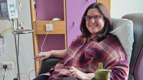 A woman with shoulder-length brown hair and glasses is smiling and sitting in a chair. She has a cannula in her hand which is wired into a machine. She is wearing a pink, purple and white plaid top.