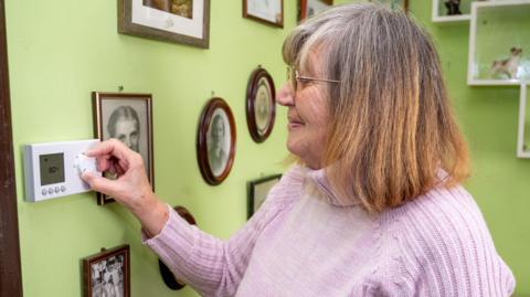 Barbara Rickard pictured in a purple sweater adjusting a dial on her thermostat on a green wall full of black and white pictures