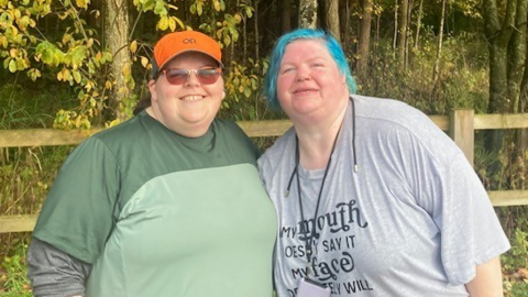 Claire Wood wears a red cap, sunglasses and a green T-shirt as she stands next to Clare Birnie who has blue hair and is wearing a grey T-shirt. 