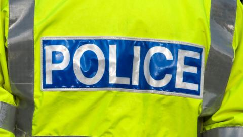 The back of a police officer's yellow high-vis jacket. There is a blue band at the back with "POLICE" written on it in white text.